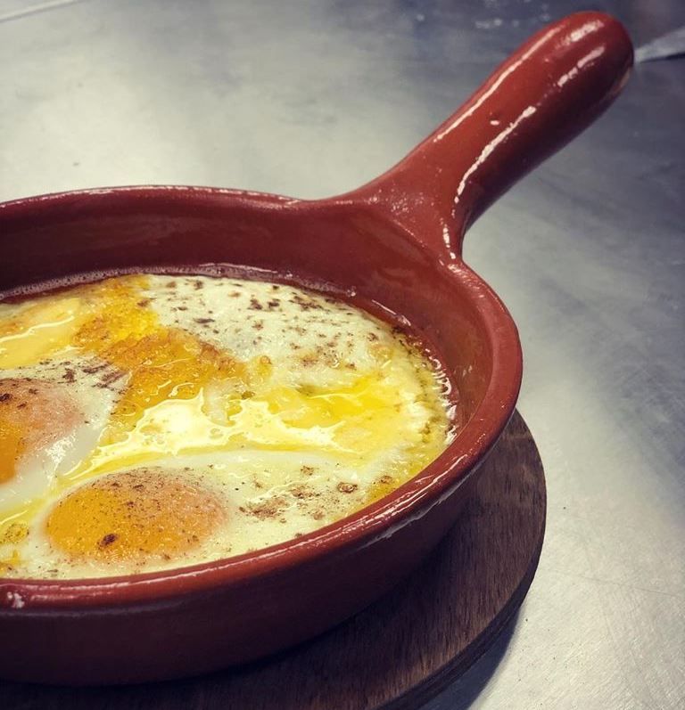 Baked eggs in a brown ceramic skillet on a wooden coaster.