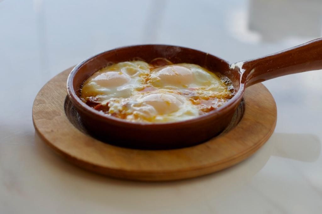 Baked eggs in a brown skillet on a wooden plate, set on a white table.