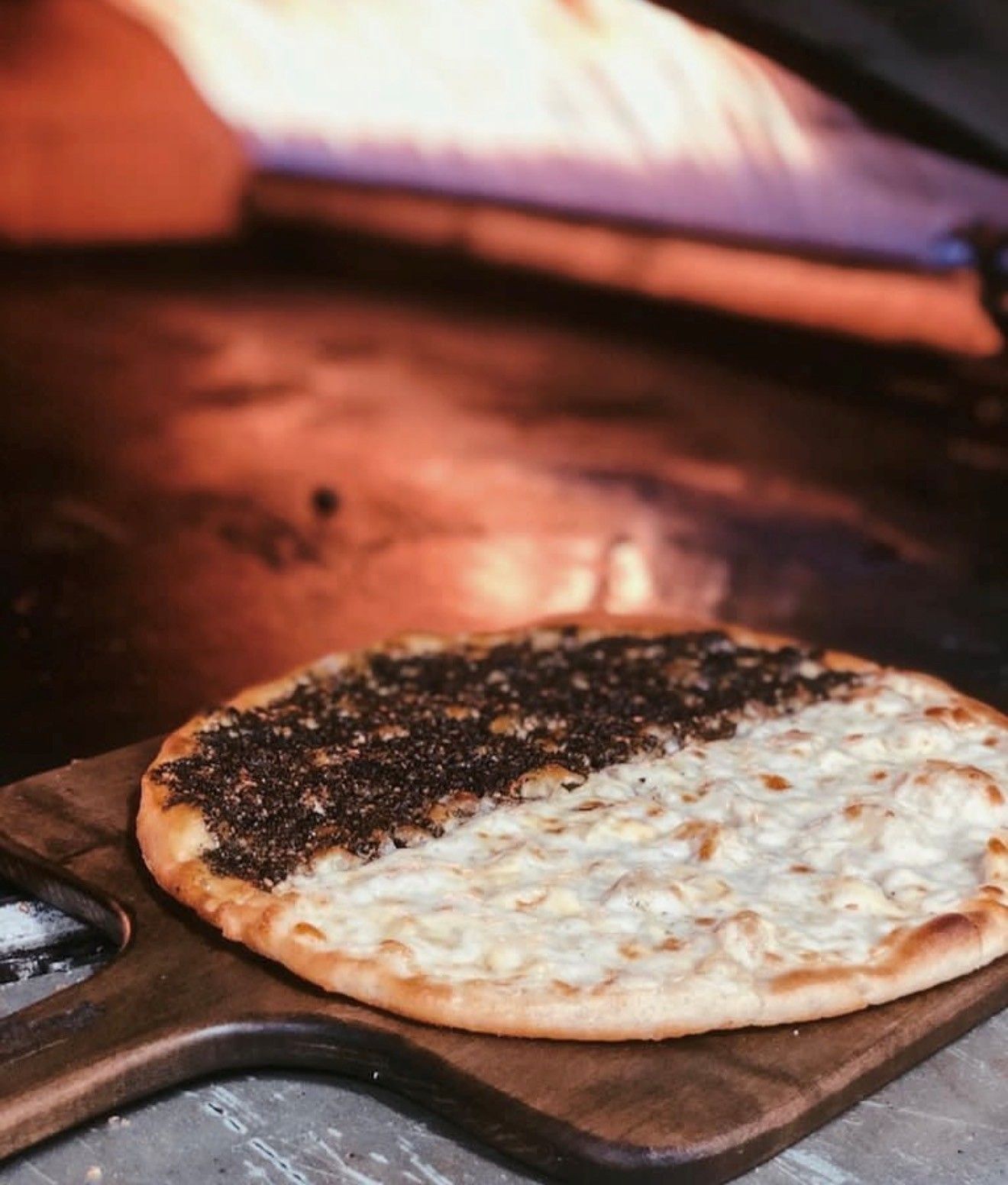 Flatbread, half za'atar, half cheese, on a wooden board in front of a wood-fired oven.