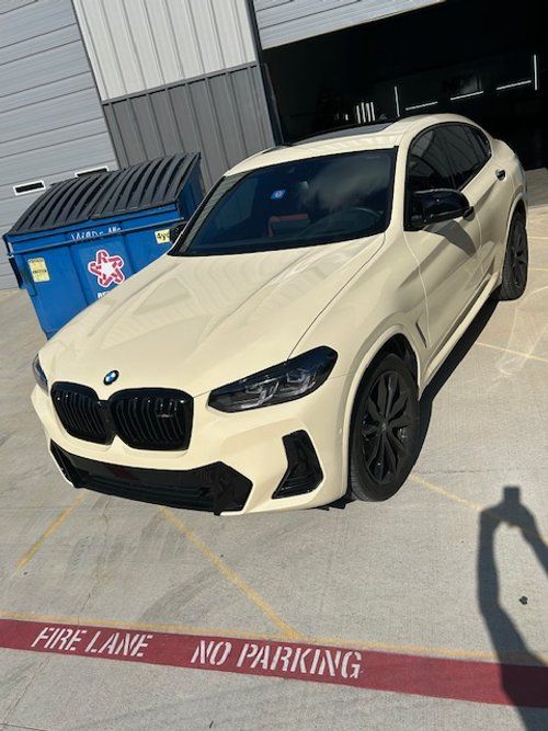A white bmw is parked in a parking lot next to a dumpster.