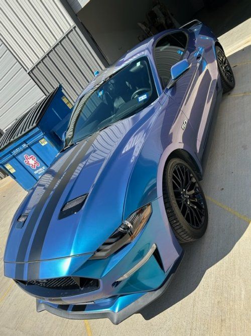 A blue mustang is parked in a parking lot next to a dumpster.