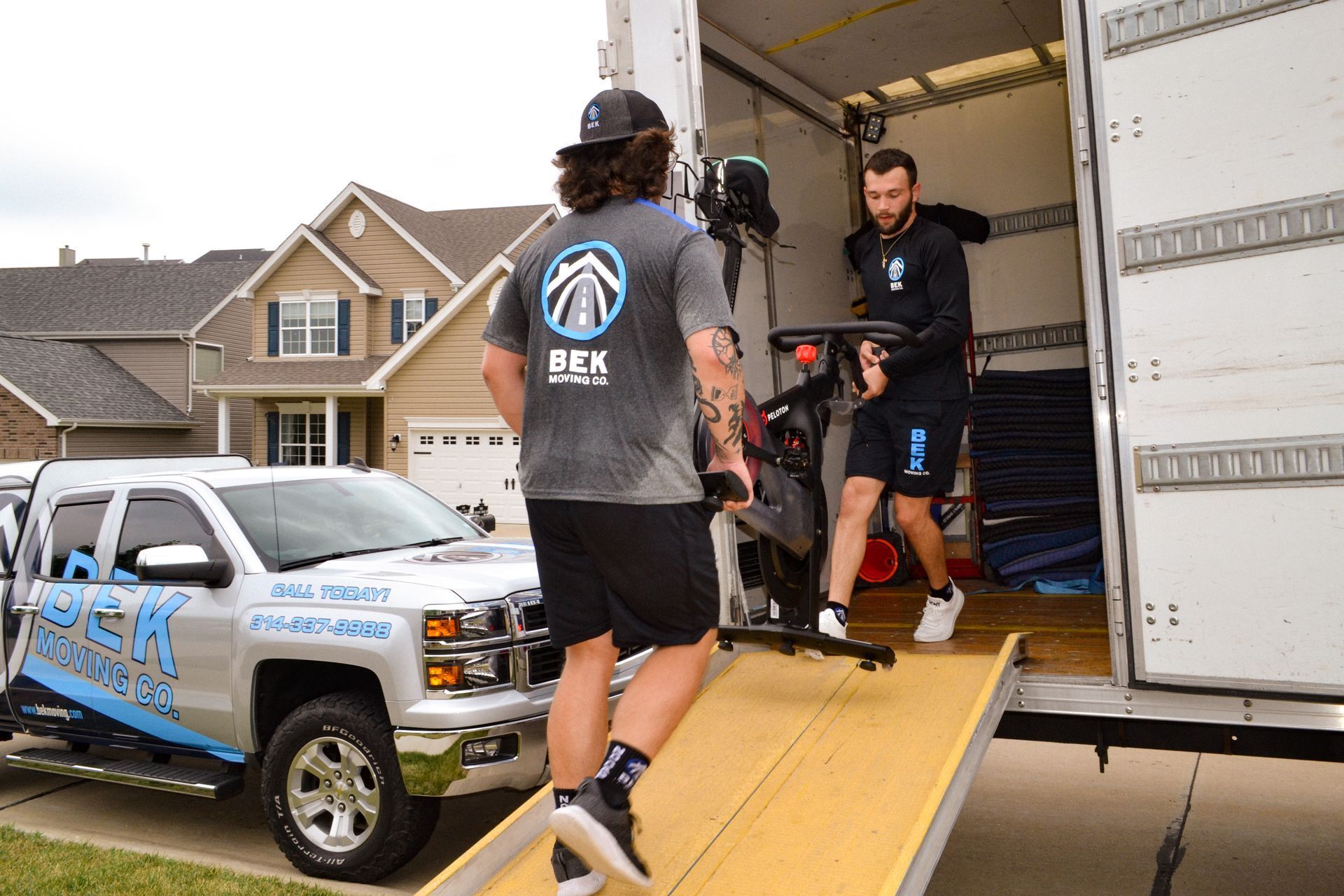 People loading gear into a truck via a ramp outside a house during a moving or event setup.