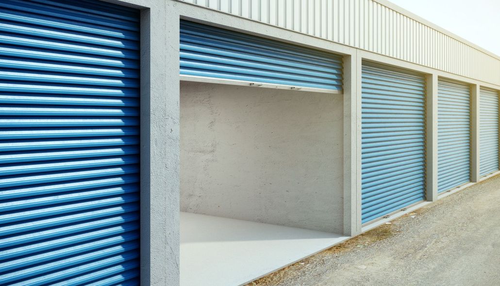 Blue roll-up storage unit doors, one open to an empty concrete bay in a row of units.