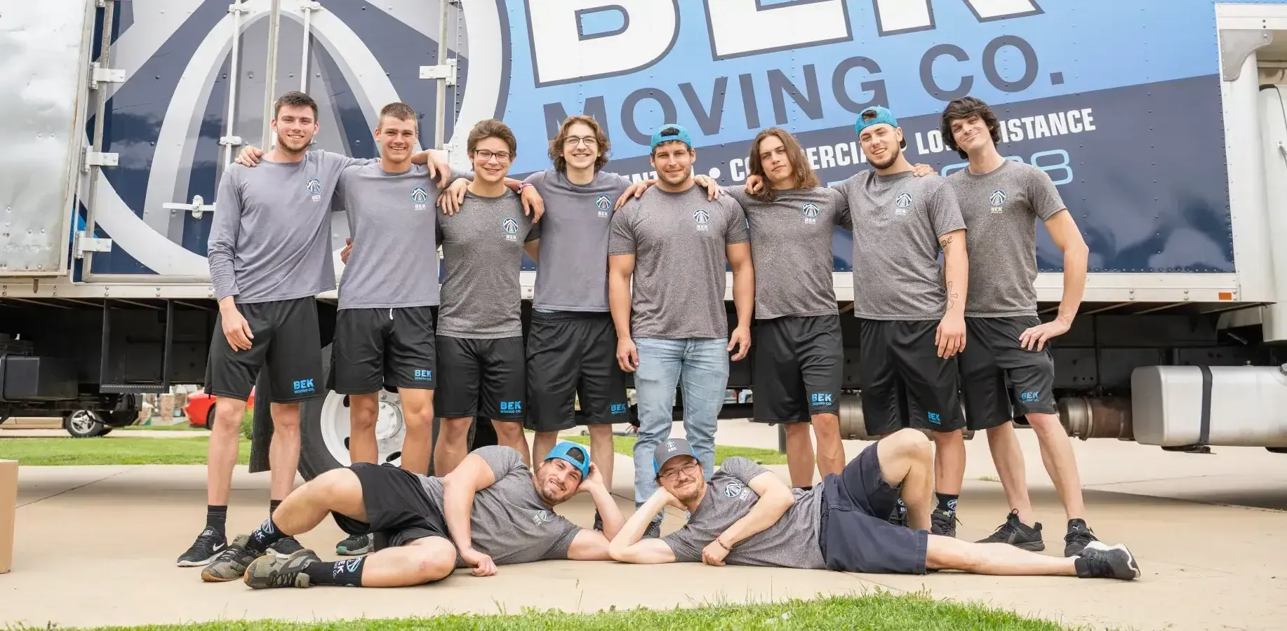 Movers posing in front of a blue moving truck, with one person lying on the ground in front.