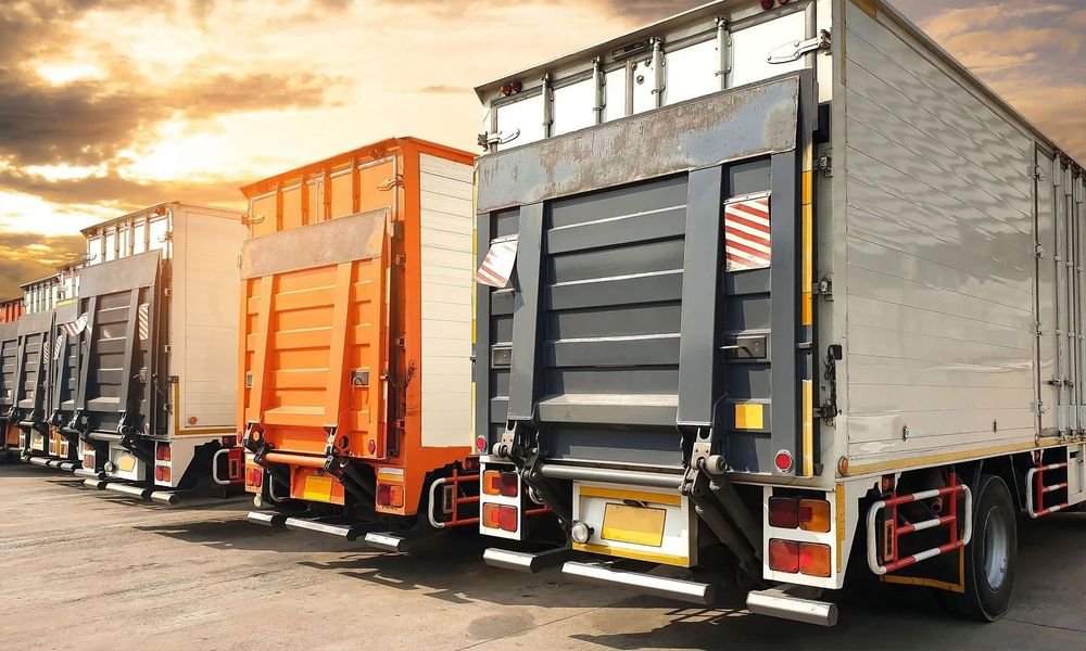 Row of semi-trucks parked in a lot, some with doors open, under a cloudy sky.