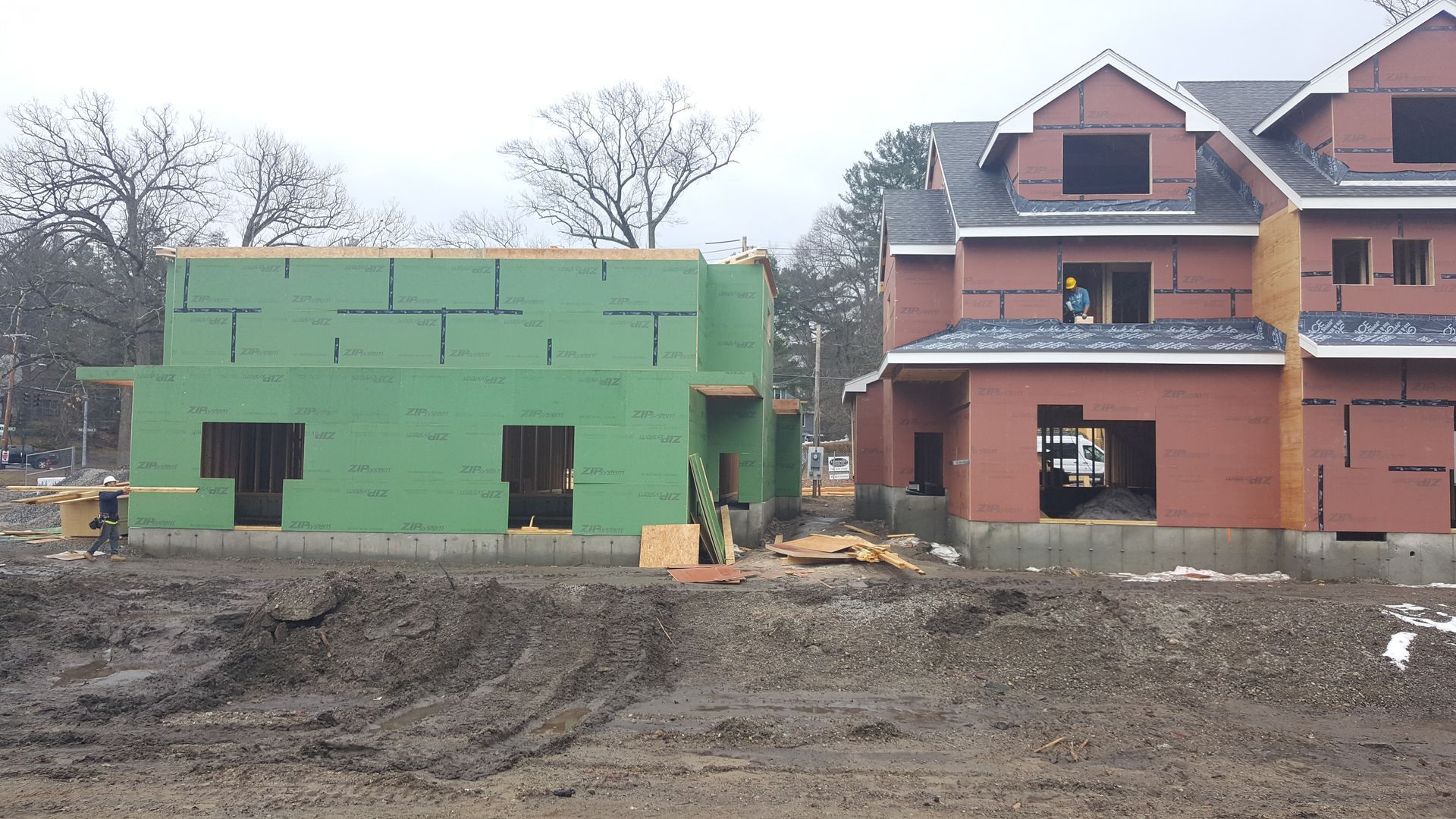 Two houses under construction, one green and one brick, on a muddy lot.