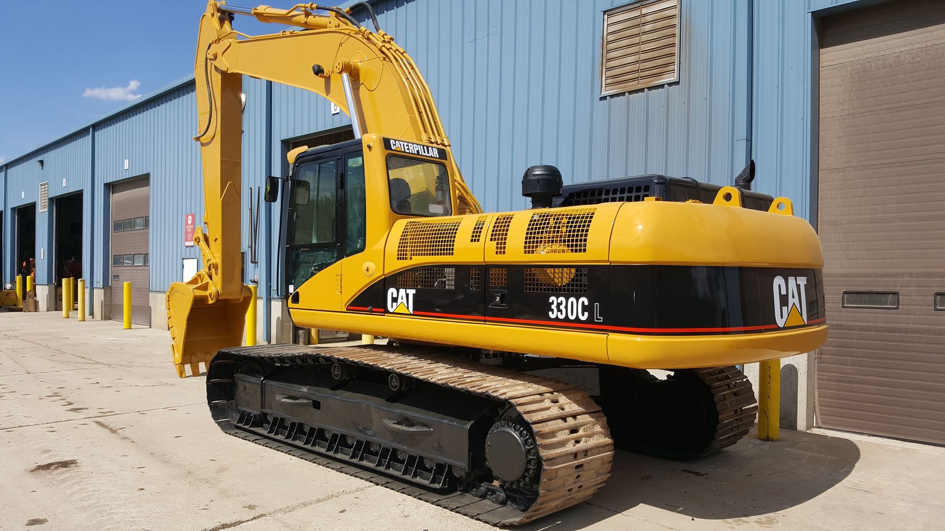Yellow CAT tracked excavator parked beside a blue warehouse on a sunny day