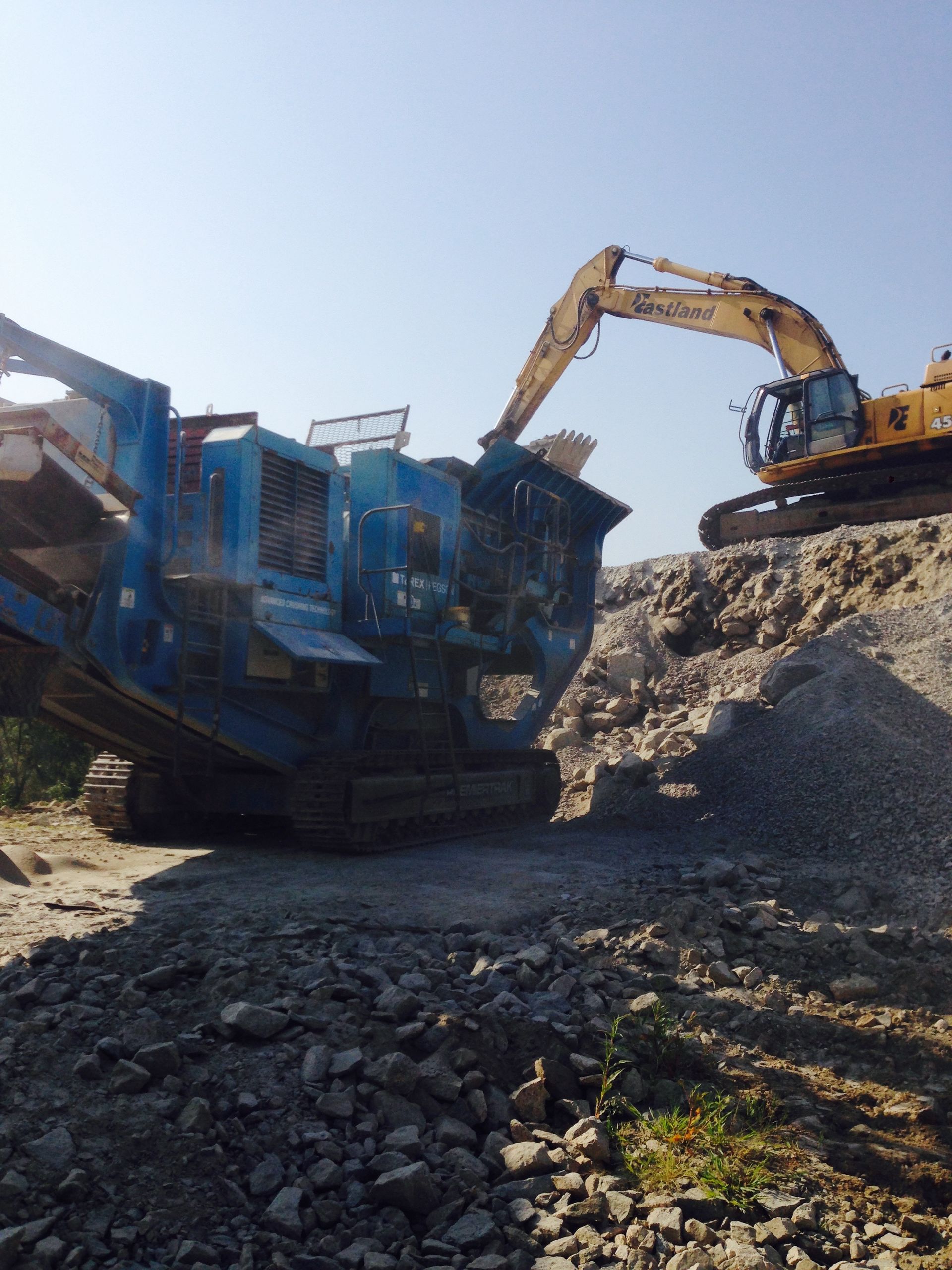 Excavator loading rock into a blue crusher at a construction site under clear sky