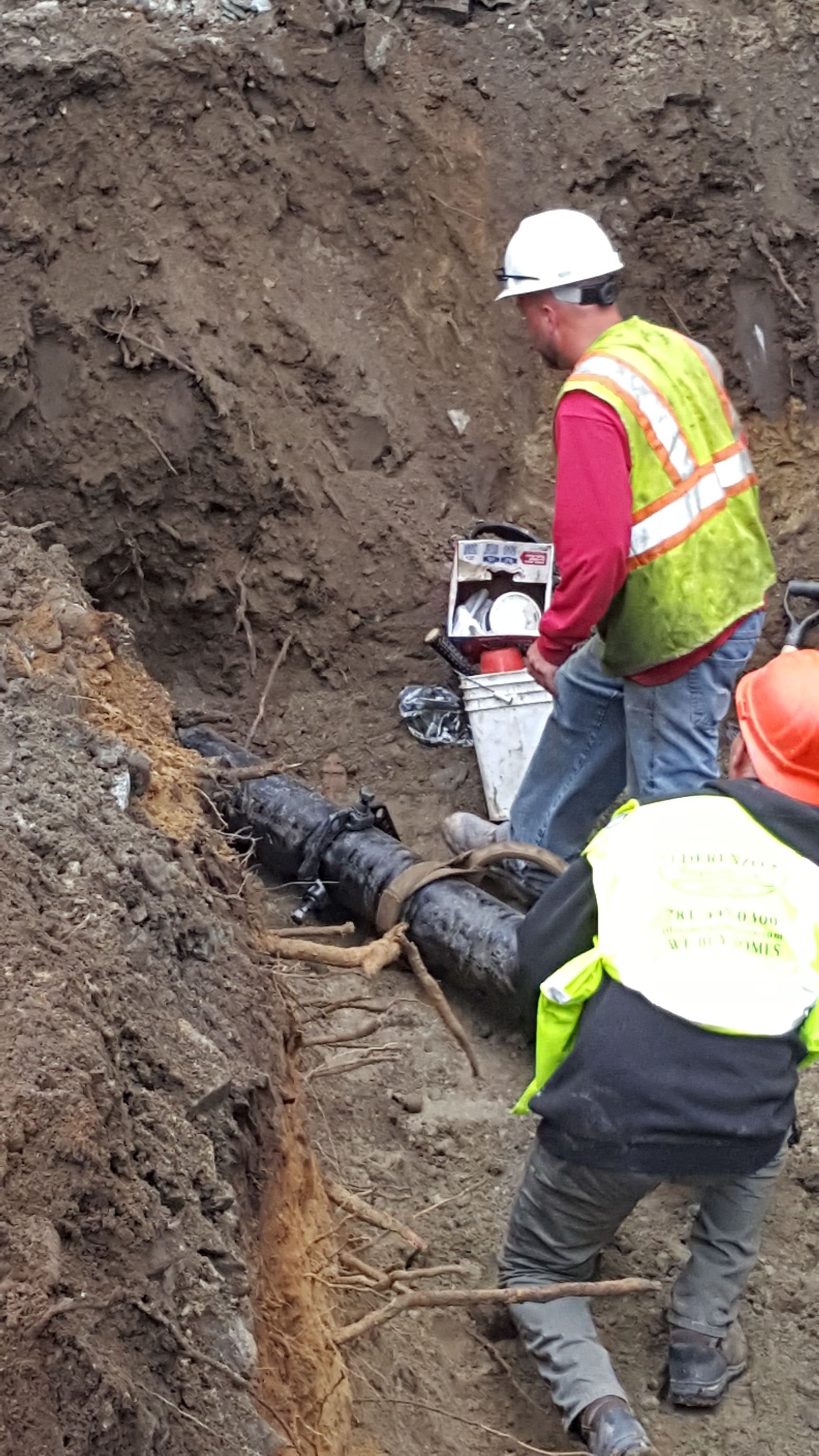 Workers in hard hats inspect a damaged underground pipe in a muddy trench.