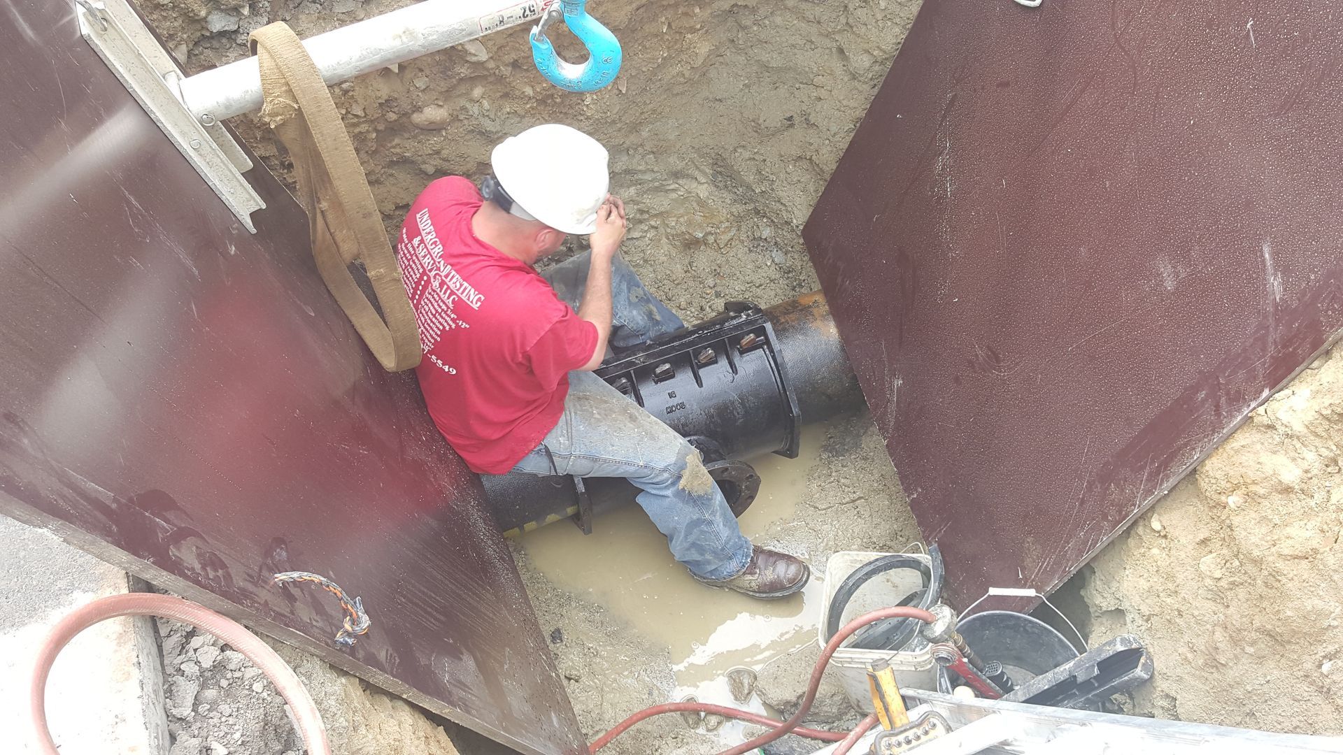 Worker in red shirt and white hard hat inspecting a pipe inside a narrow trench.