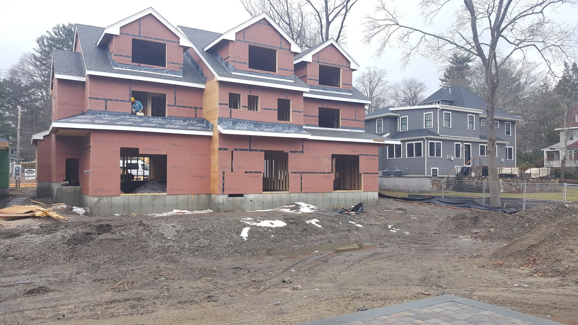 Partially built red-brick house on a muddy lot with bare trees and neighboring homes in the background