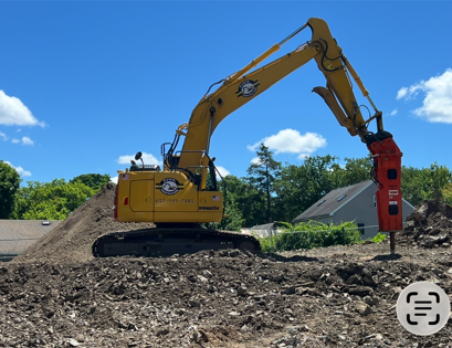 Yellow excavator with a red hydraulic breaker on a dirt site under a blue sky