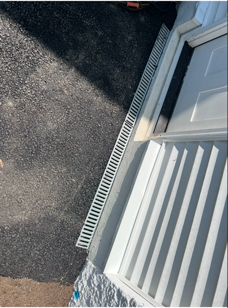 White door threshold with metal drain grate beside dark asphalt and a small section of white siding