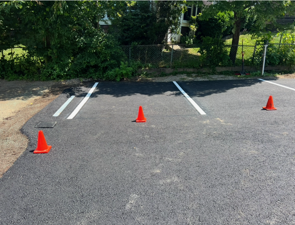 Parking lot with orange traffic cones marking spaces on asphalt near a fence and trees.