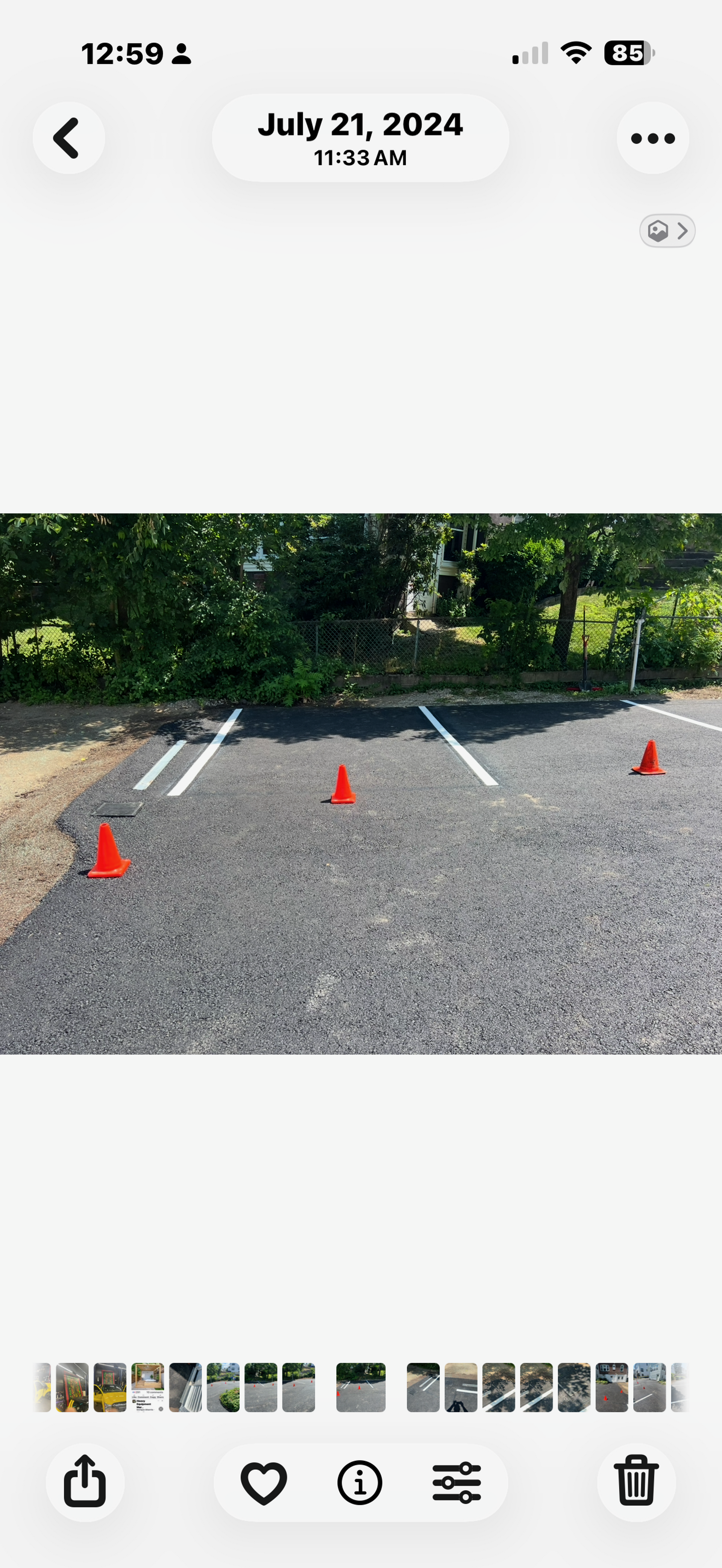 Gravel lot with red cones marking spots beside a green hedge and trees, viewed in a phone gallery screenshot