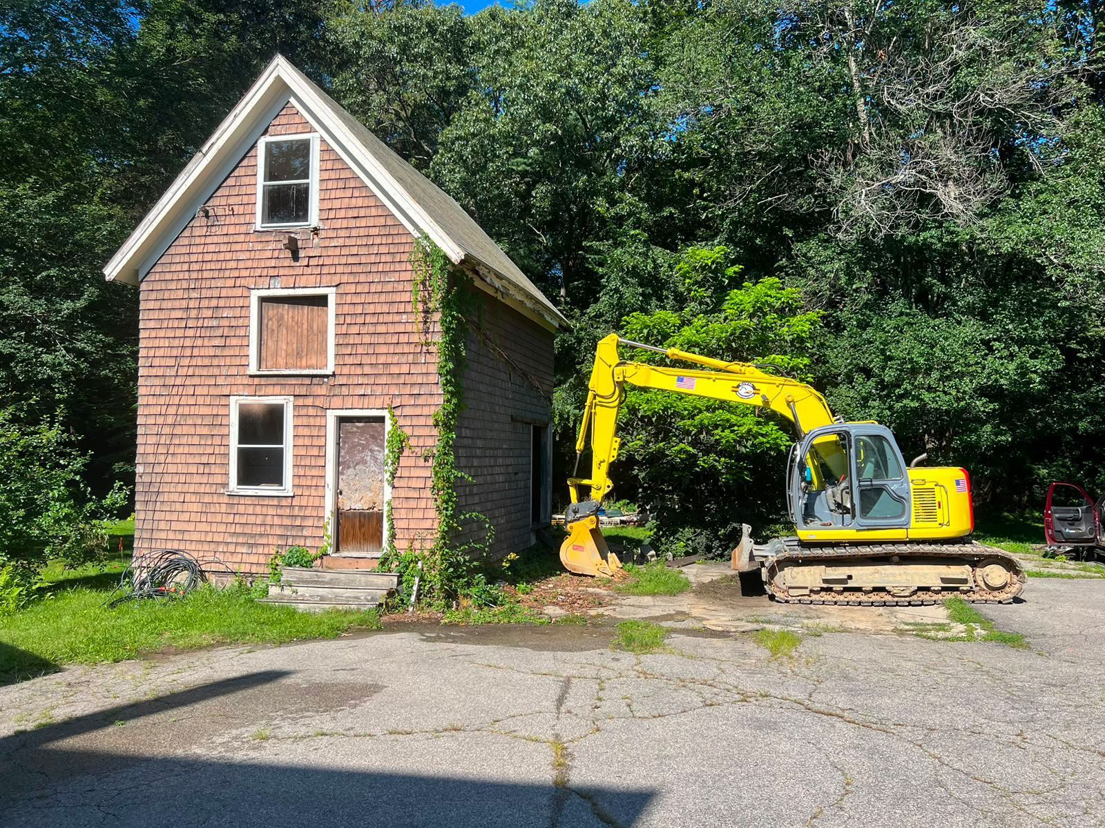 Yellow excavator beside a small red brick house with a boarded door and windows in a grassy lot