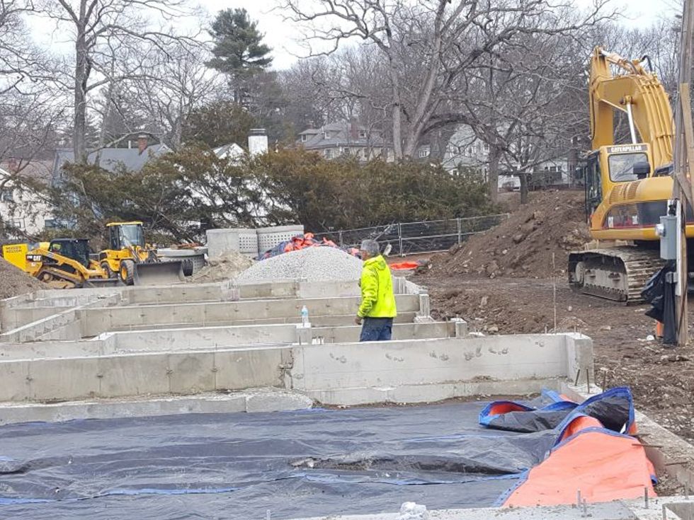 Construction site with concrete blocks, excavator, and worker in a neon jacket outdoors.
