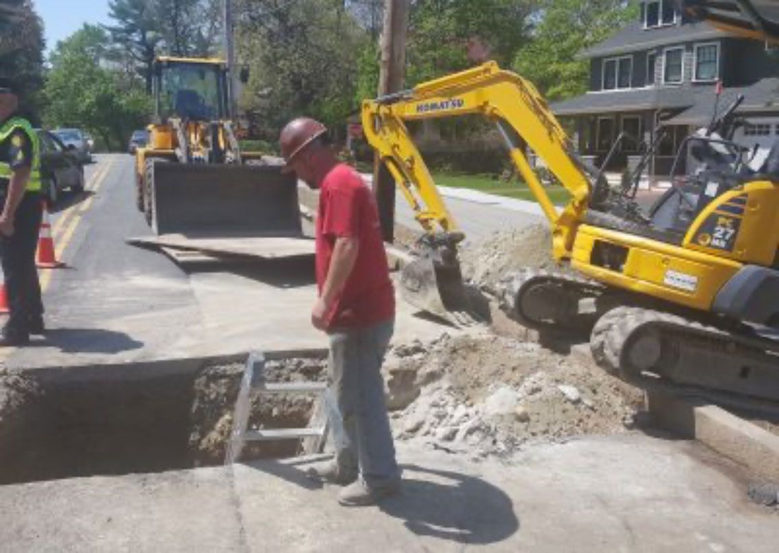 Roadwork crew with excavators repairing a street trench beside houses and a worker in a red shirt.