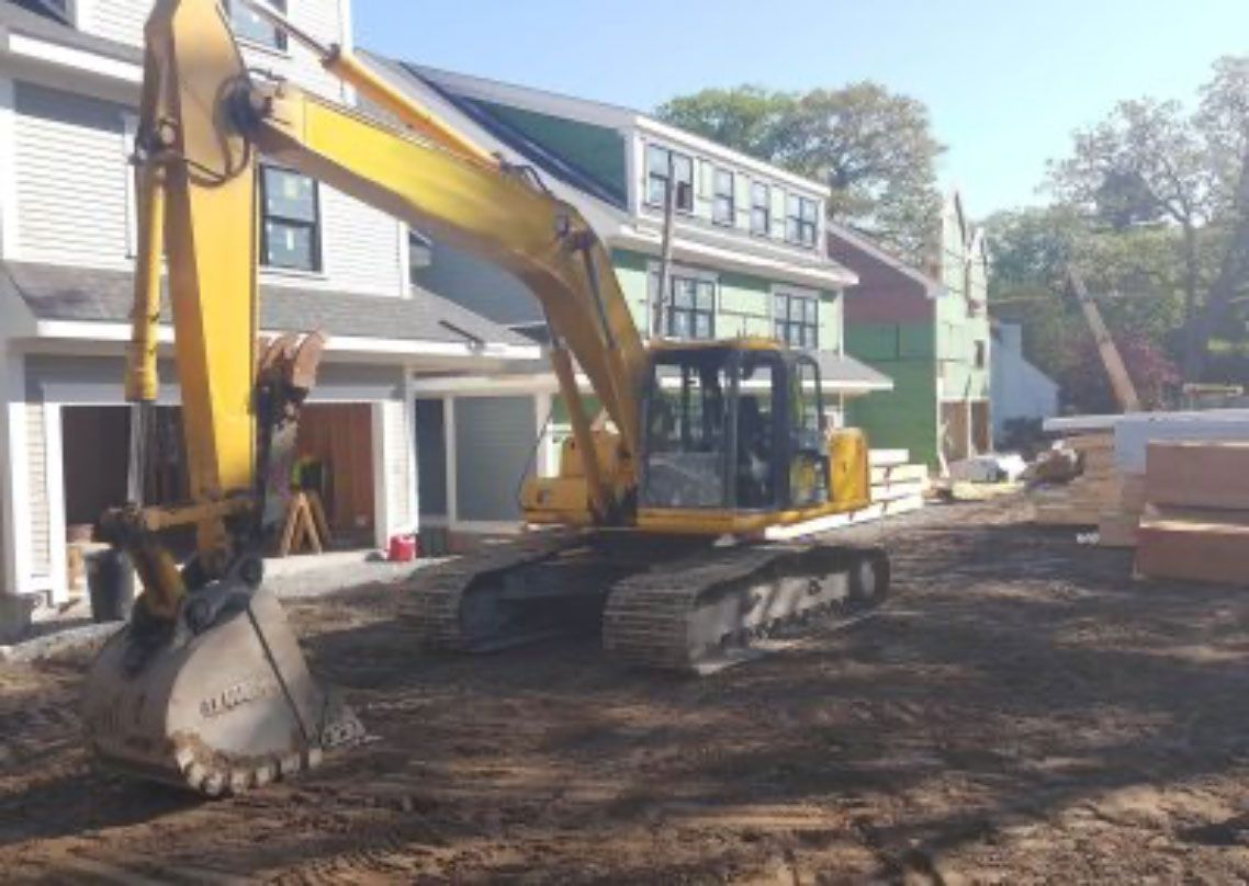 Yellow excavator on a dirt lot beside houses during construction