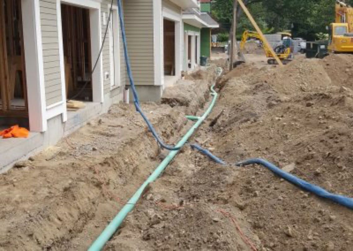 Trenches dug beside townhouses with exposed utility pipes and construction equipment in a muddy worksite.