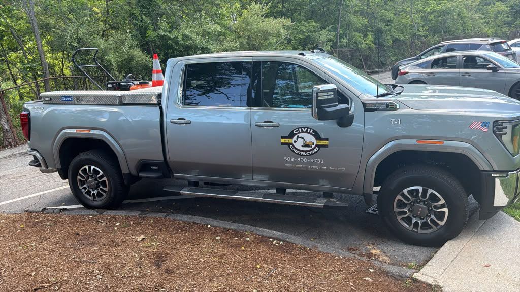 Silver pickup truck with logo parked on a wet driveway beside trees and other cars