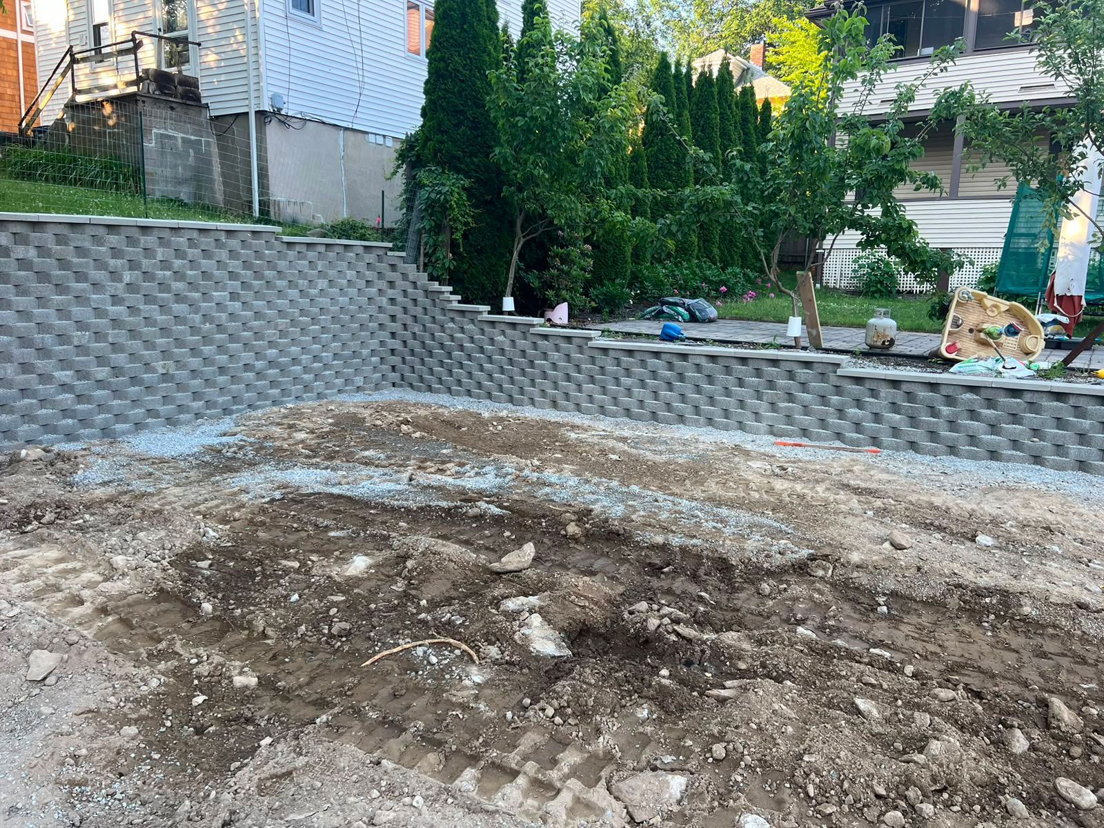 Rough dirt excavation area beside a gray retaining wall in a backyard, with trees and a house in the background