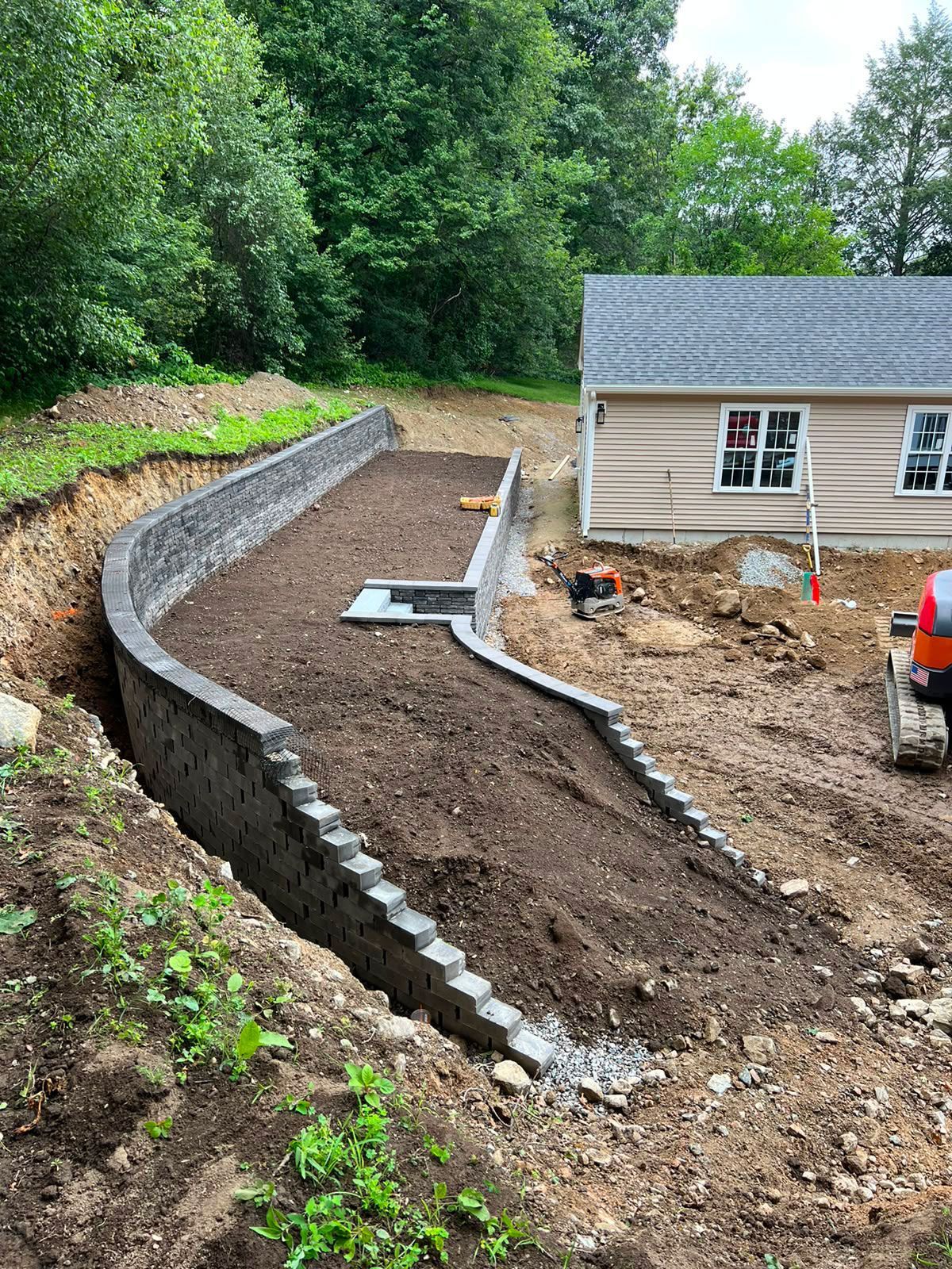 Freshly graded backyard beside a house, with a curved stone retaining wall and dirt landscaping under construction