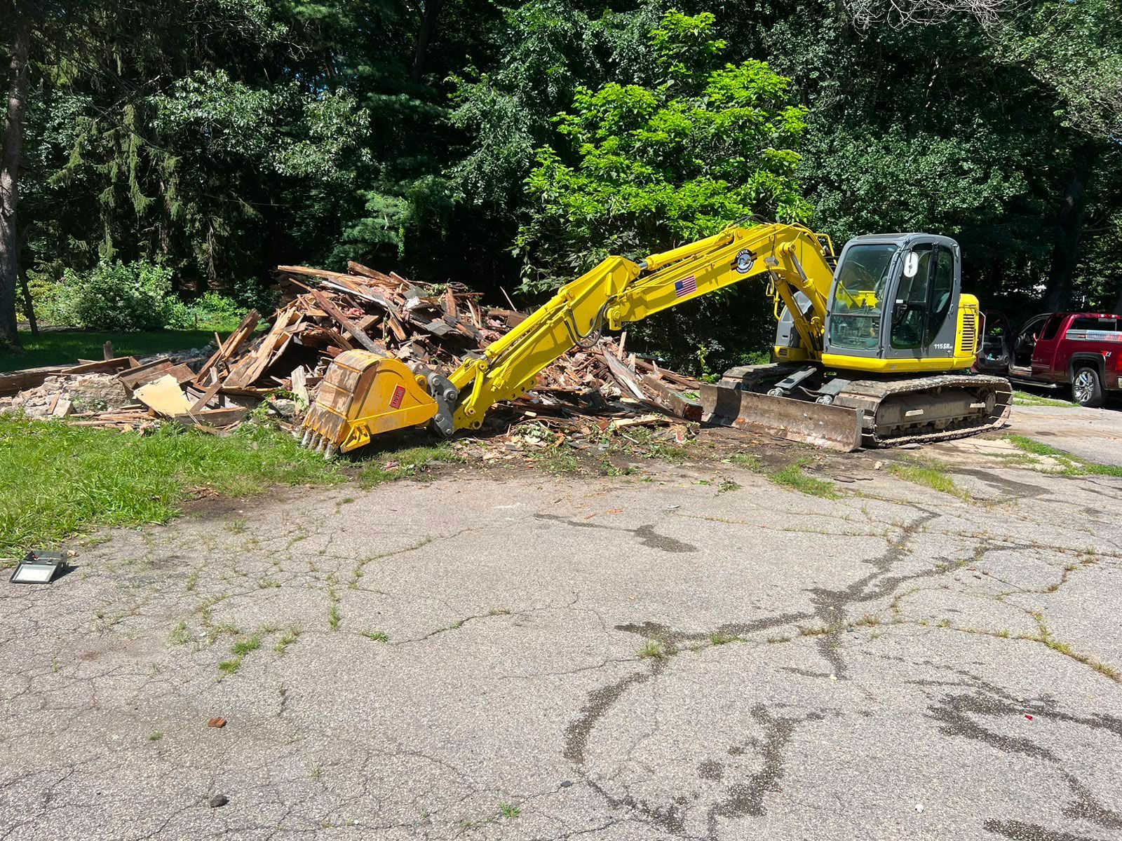 Yellow excavator clearing a debris pile beside a cracked paved lot and trees