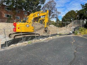 Yellow excavator clearing dirt beside a brick building and chain-link fence along a paved road.
