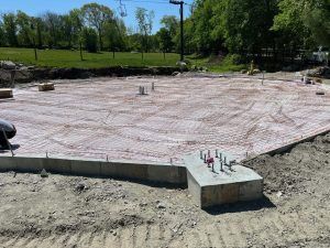 Concrete foundation slab under construction in a grassy park area, with rebar and utility pipes visible.