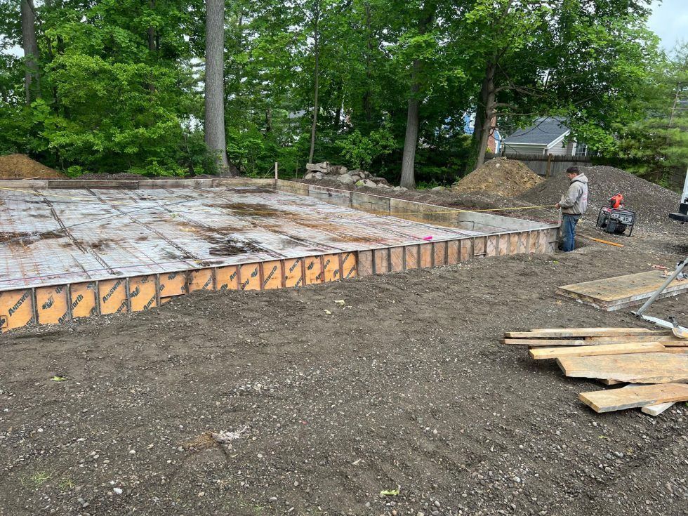 Construction site with a large foundation area, workers, gravel, and trees in the background.