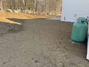Gravel yard beside a white house, with a green propane tank and trees in the background.