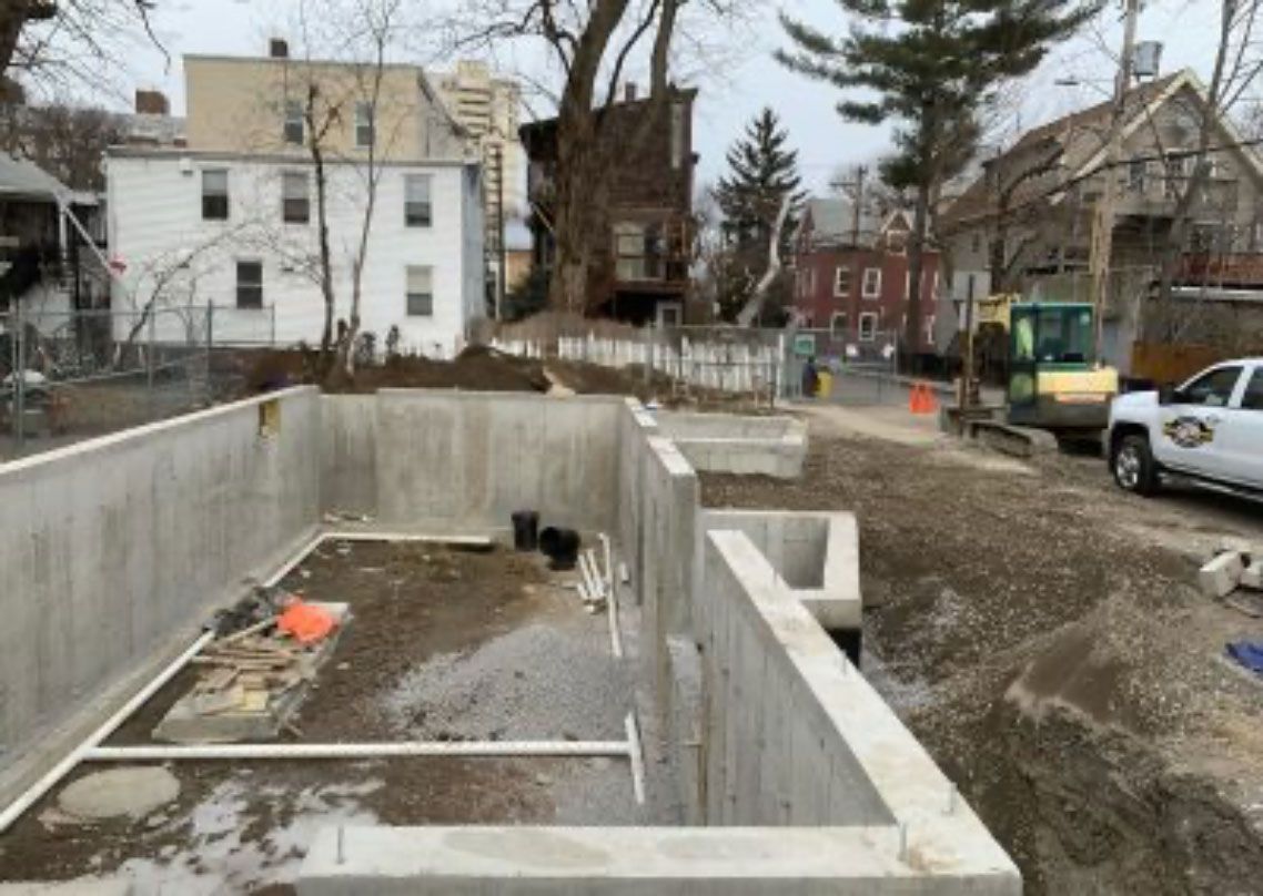 Construction site with a concrete foundation pit beside houses and parked vehicles