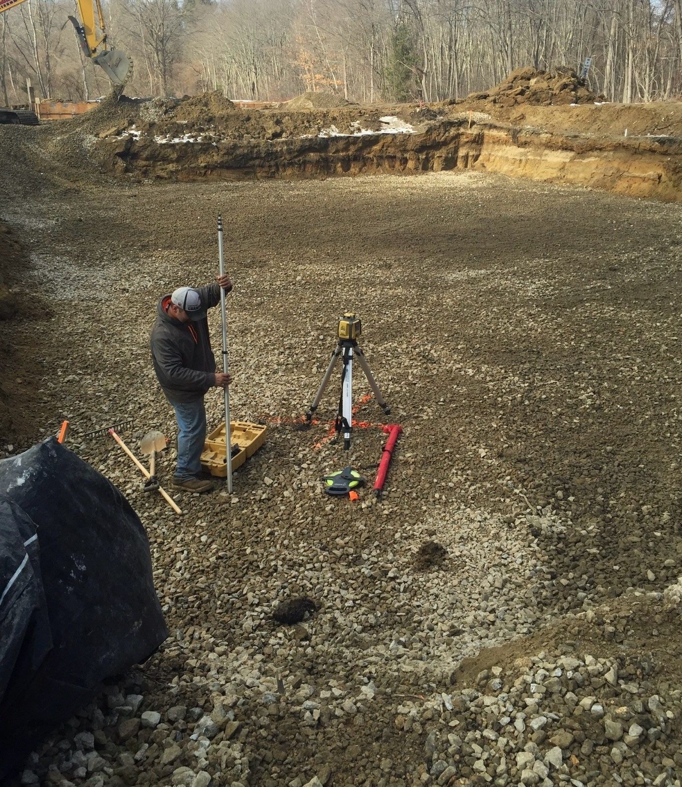 Surveyor using tripod equipment in a muddy excavation site with a wooded dirt embankment.