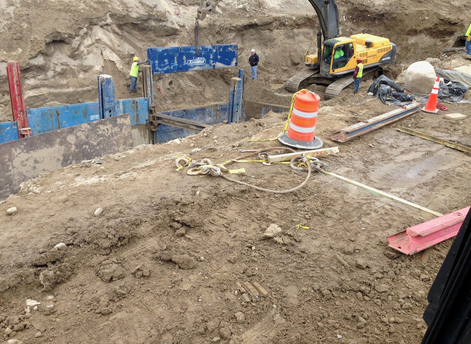 Construction site with excavator, orange safety barrels, and blue retaining wall in a dirt excavation area