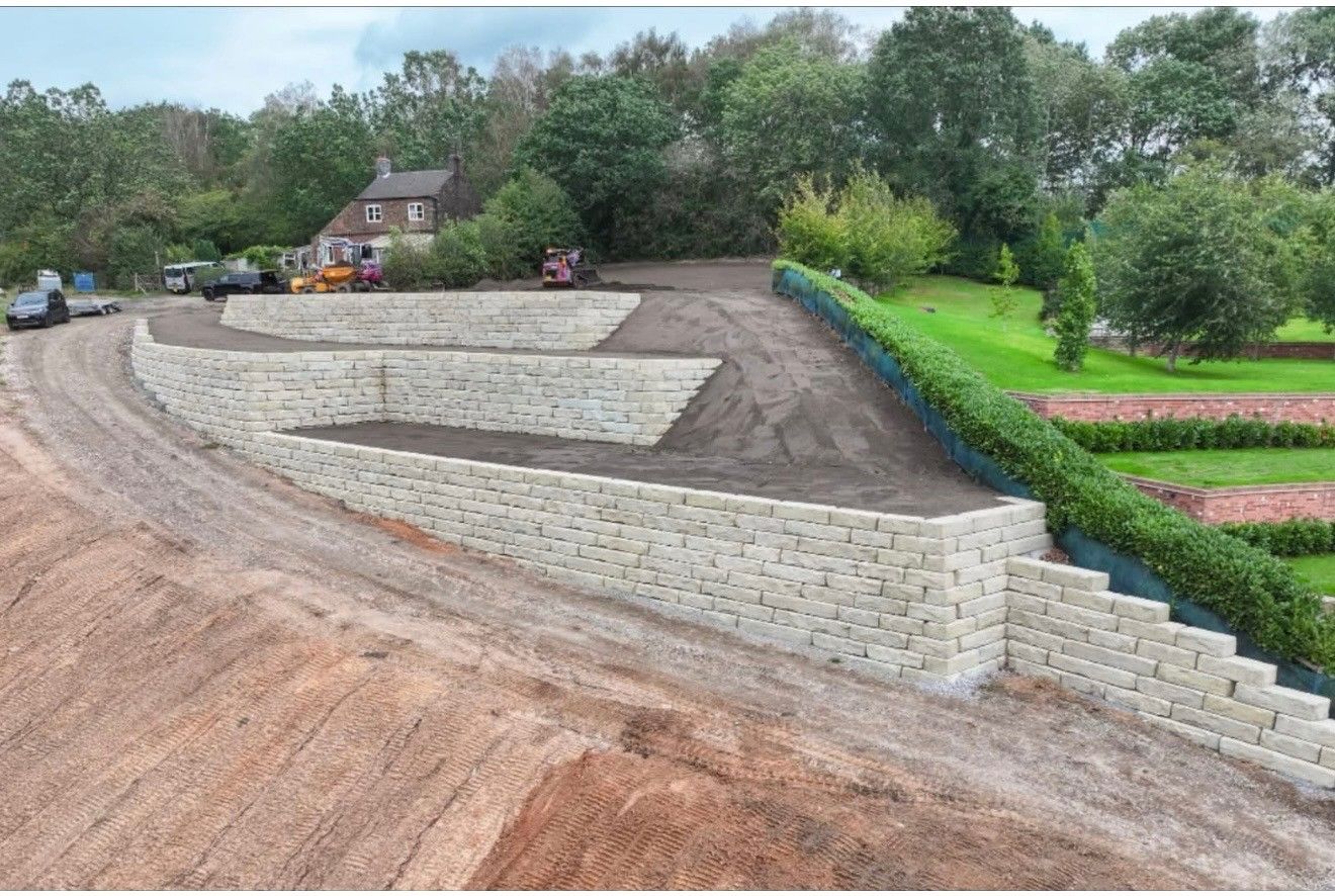 Terraced stone retaining walls on a sloped landscaped construction site with trees and houses nearby.