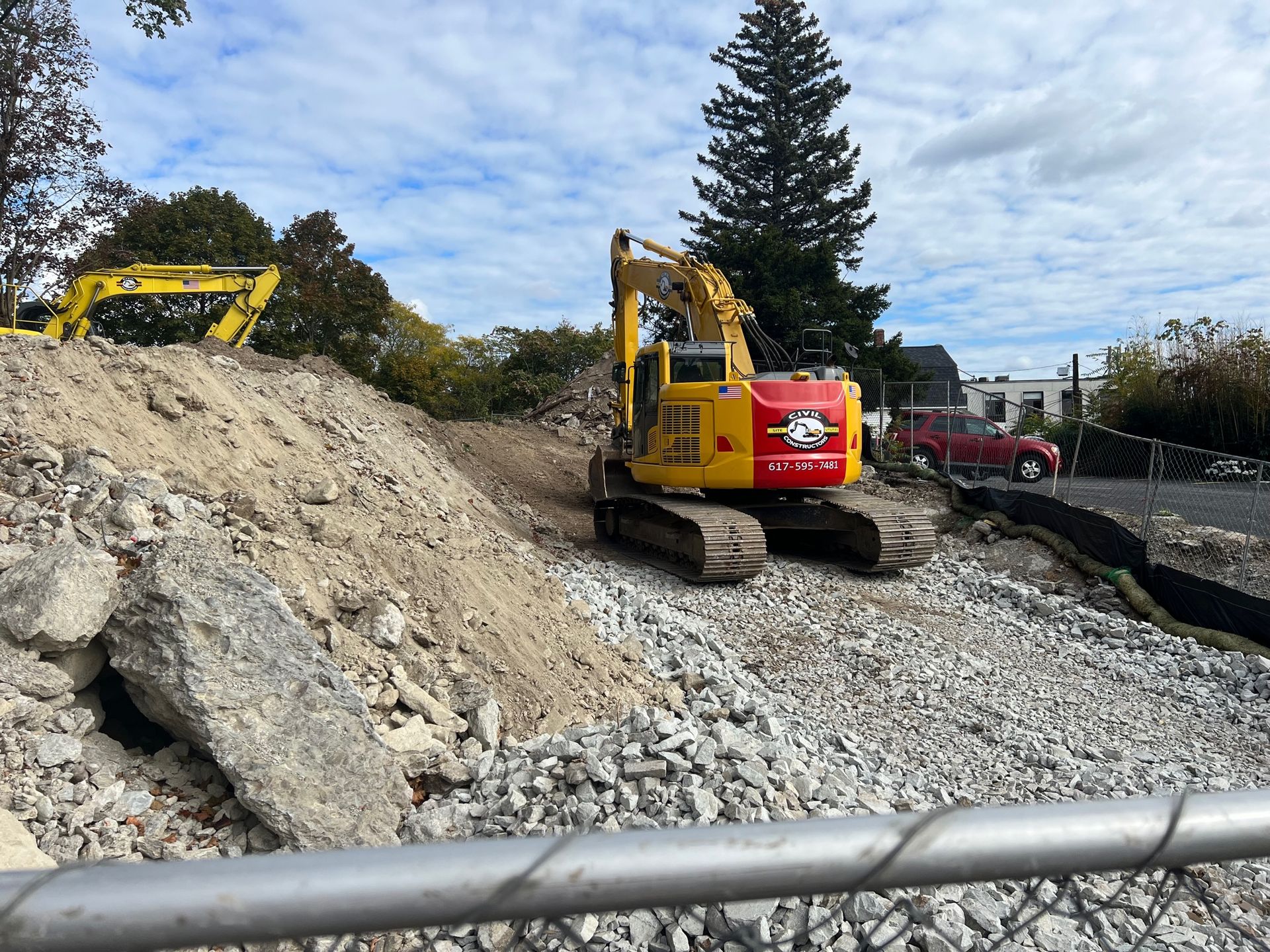 Two yellow excavators on a rocky construction site with a red truck and trees in the background