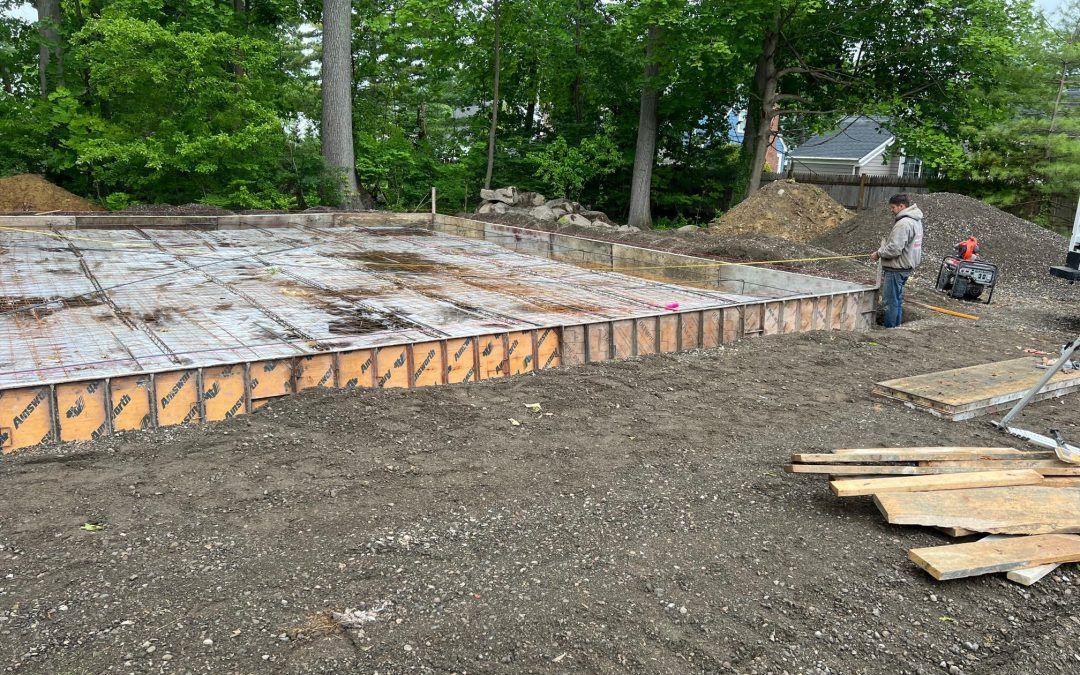 Construction site with a long stone retaining wall and workers on the right, surrounded by trees.