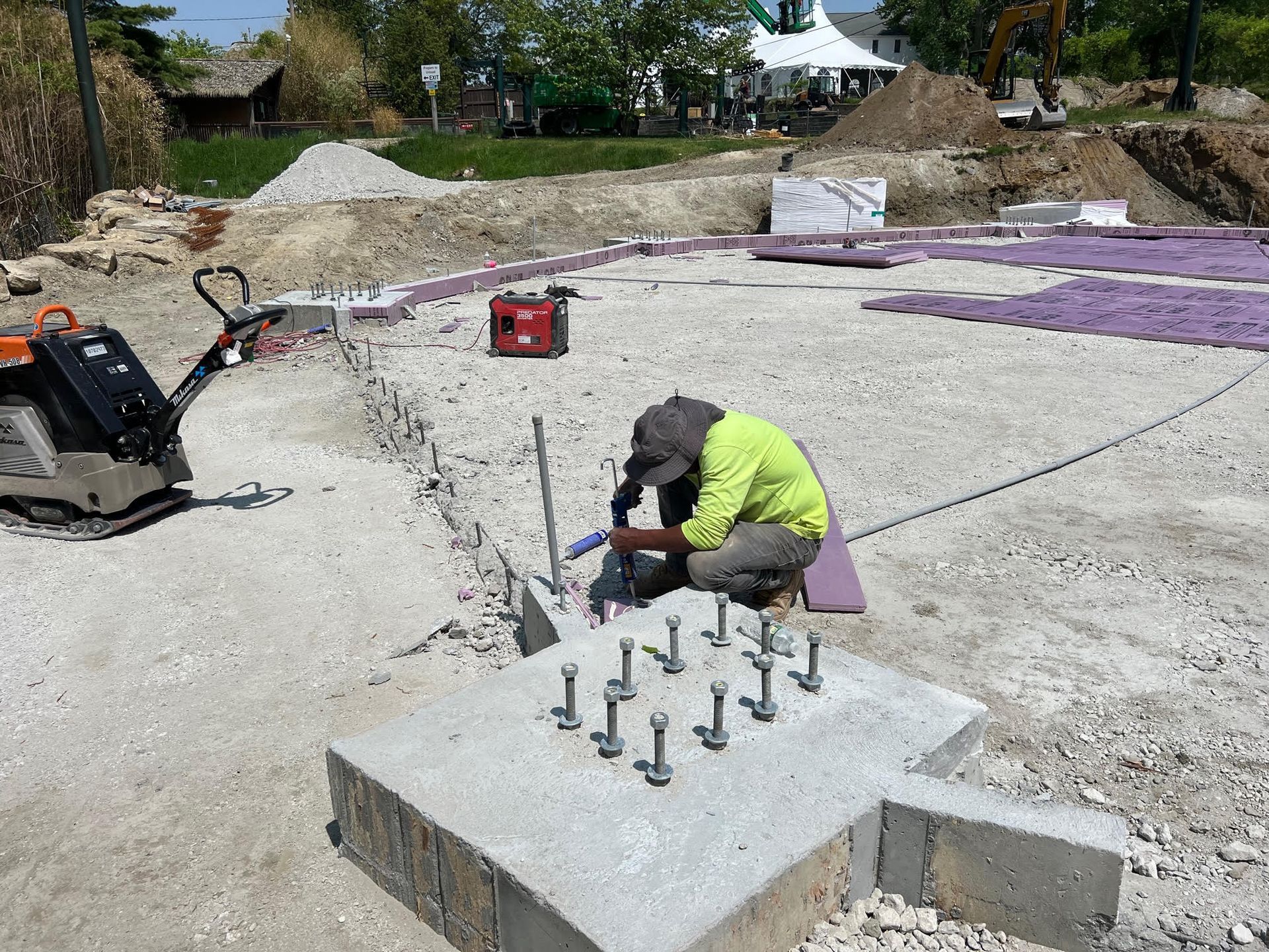 Worker kneeling on concrete footing, drilling rebar anchors at a construction site.
