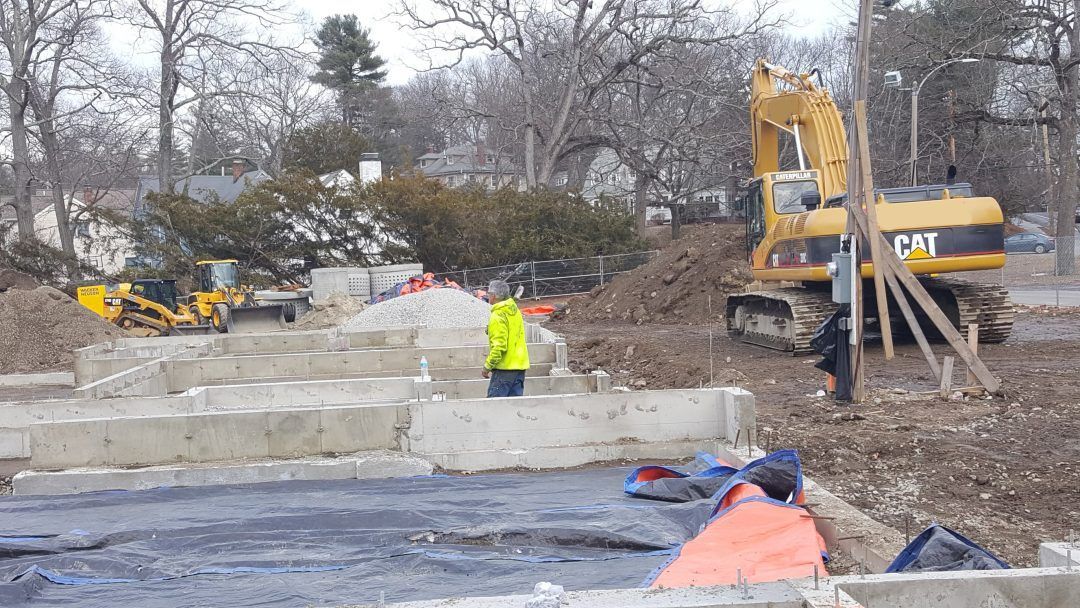 Construction site with excavator, worker in yellow vest, and concrete blocks near a wooded area