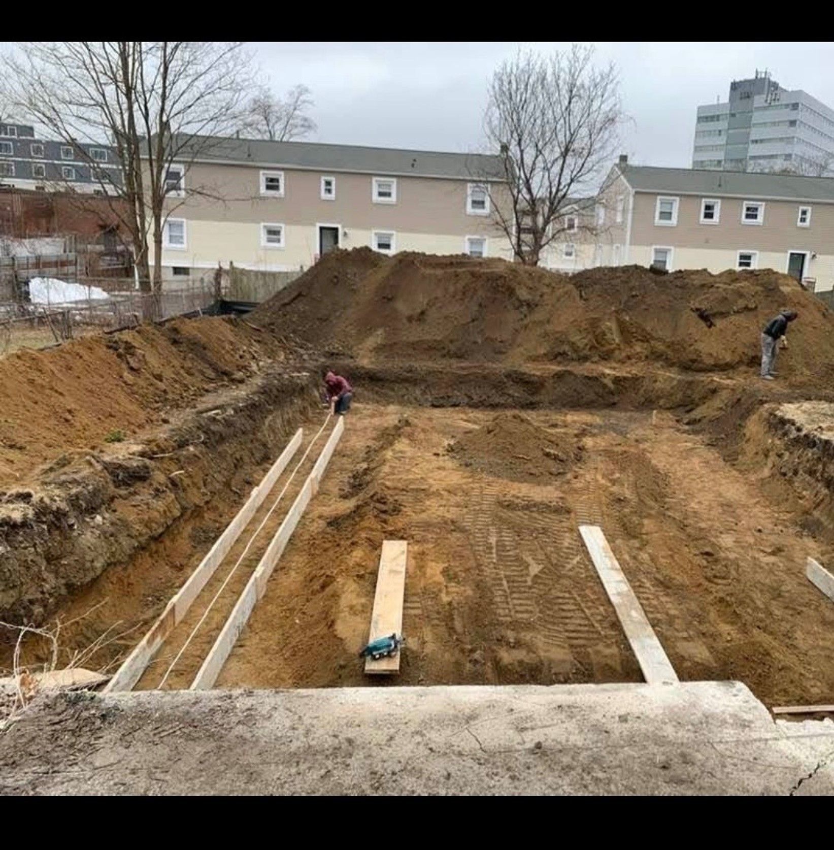 Large excavated construction pit with workers, soil piles, and apartment buildings in the background
