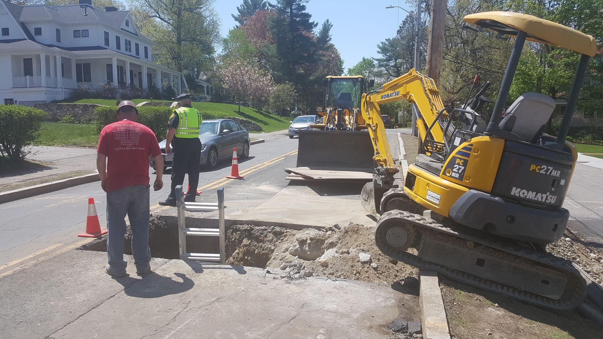 Roadwork scene with excavator, workers, and a hole cut into the street on a sunny residential road