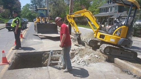 Roadwork scene with a yellow excavator, workers, and an open trench in the street