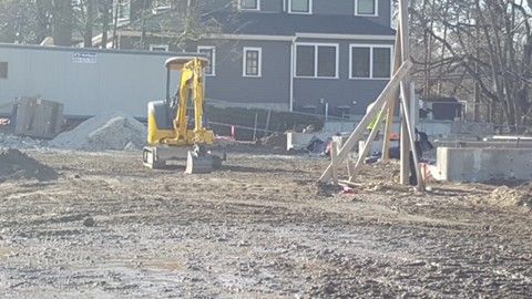 Construction site with a yellow excavator in front of a house and piles of dirt.