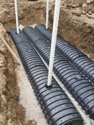 Black corrugated pipes laid in a trench beside white vertical pipes and gravel foundation work