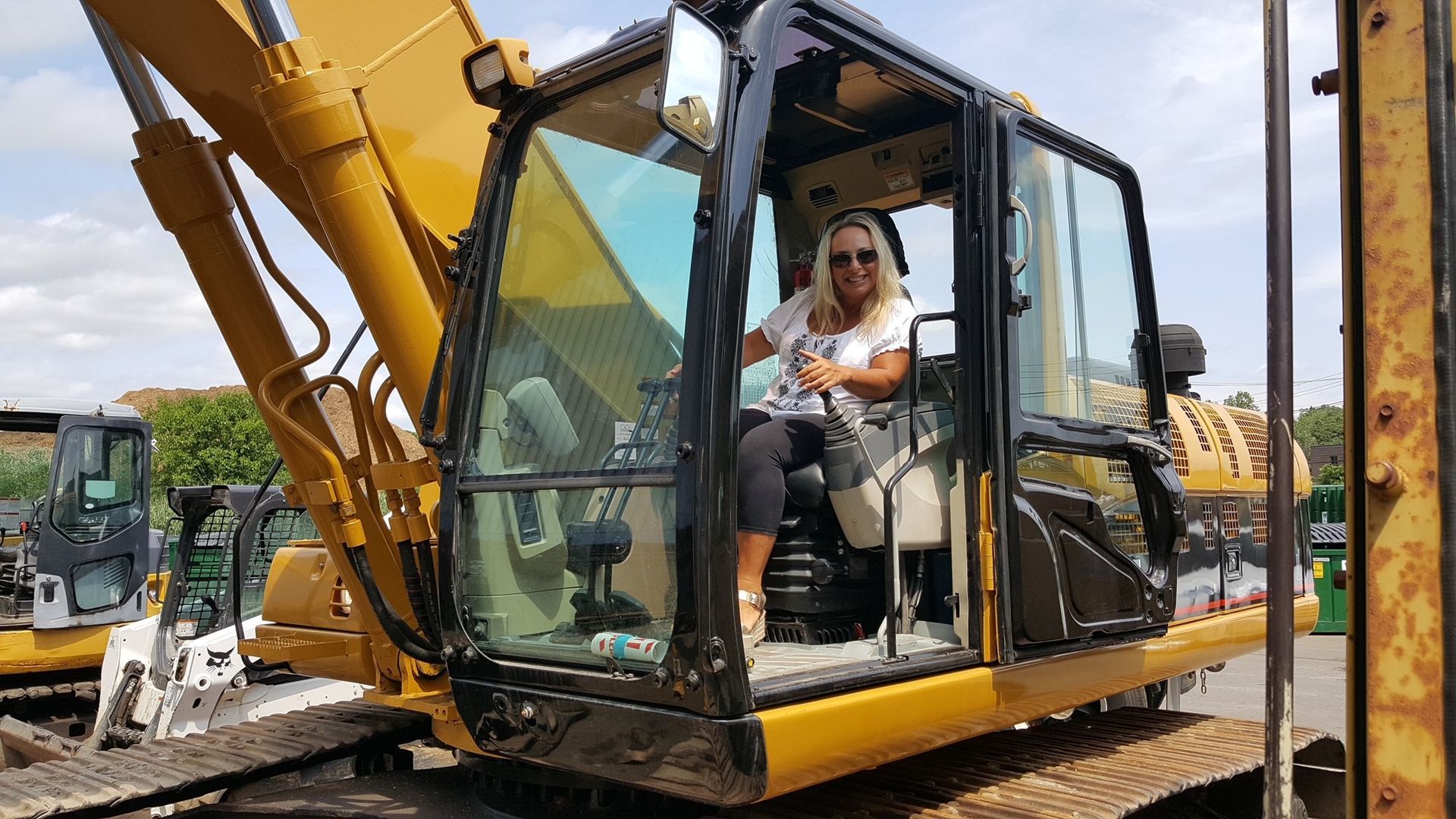 Woman seated in a yellow excavator cab at an equipment yard, smiling at the camera.
