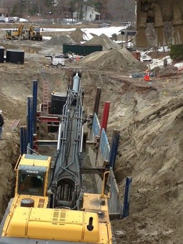 Construction site with drilling rig and yellow machinery in a deep trench, dirt piles in the background