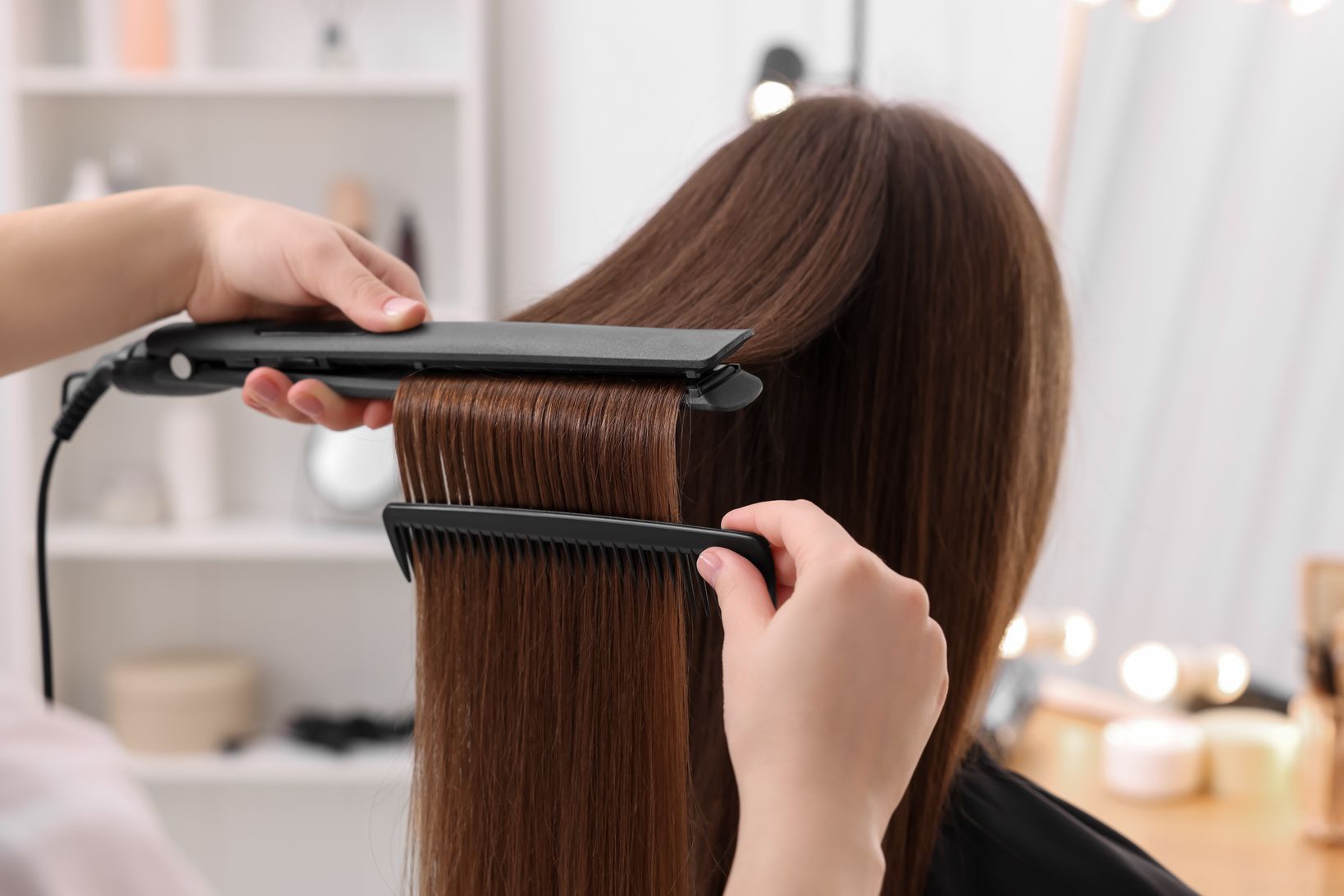 Hair stylist straightens a customer’s brown hair with a flat iron and comb in a salon.