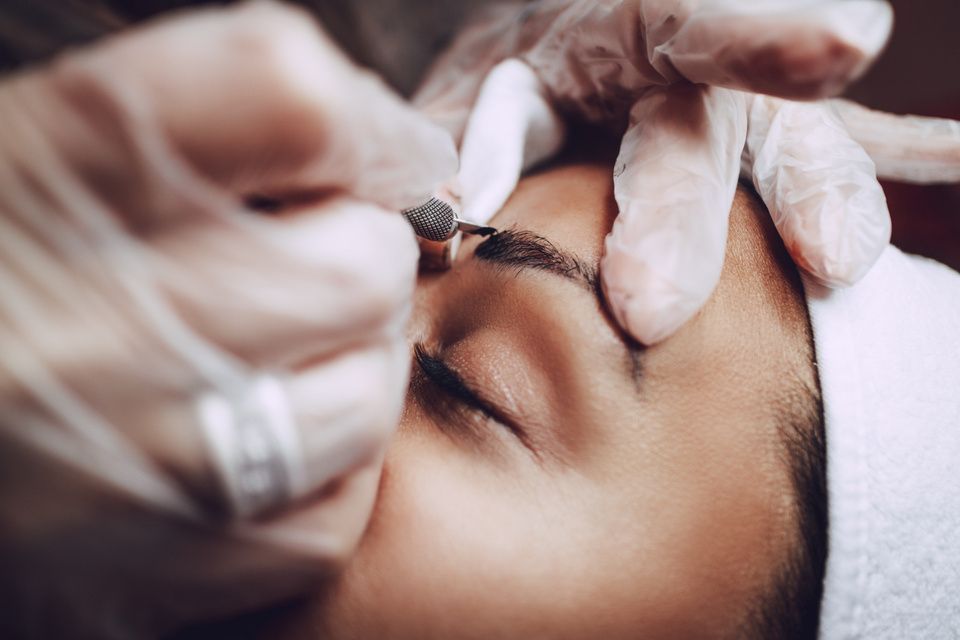 Person receiving eyebrow microblading; gloved hands, closed eyes.