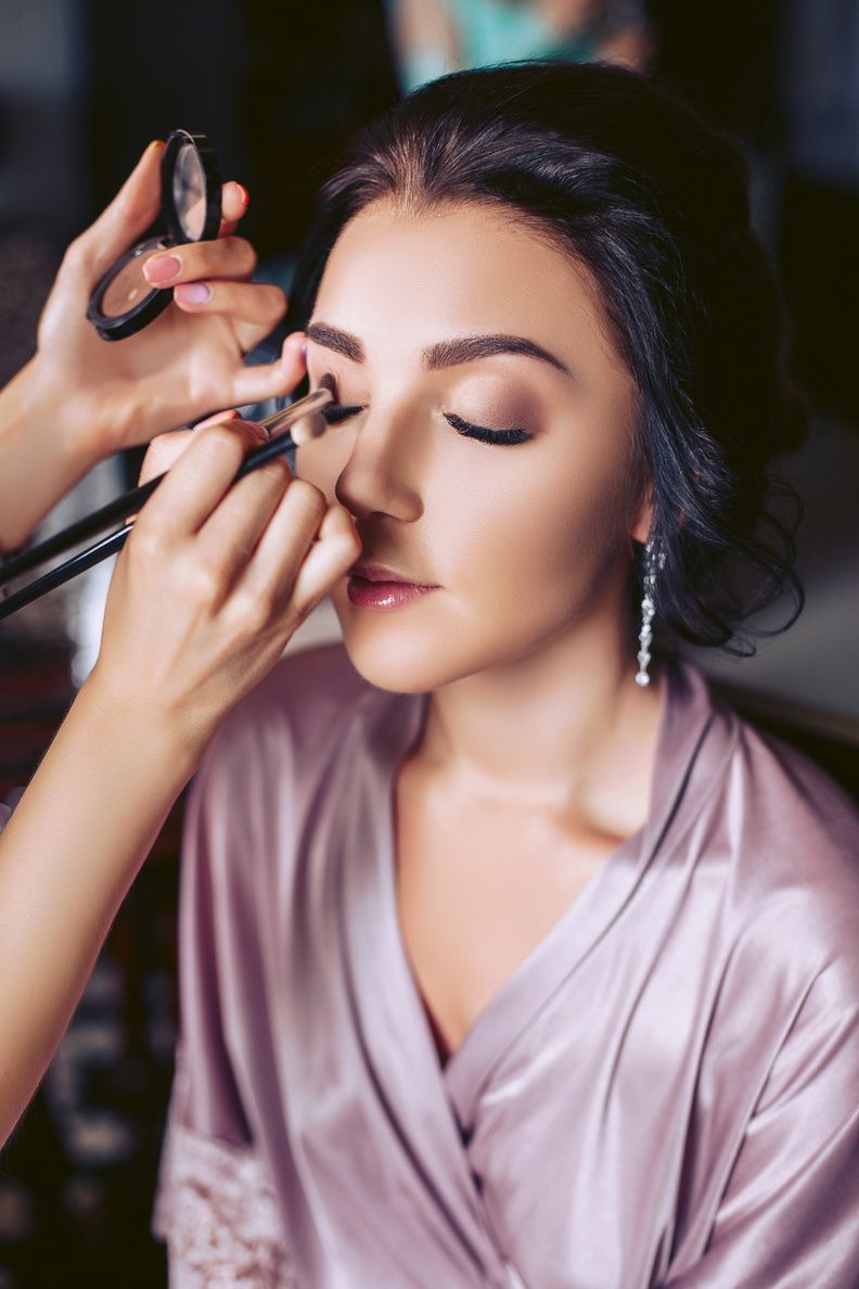 Woman having eye makeup applied, wearing a pink robe, with eyes closed.